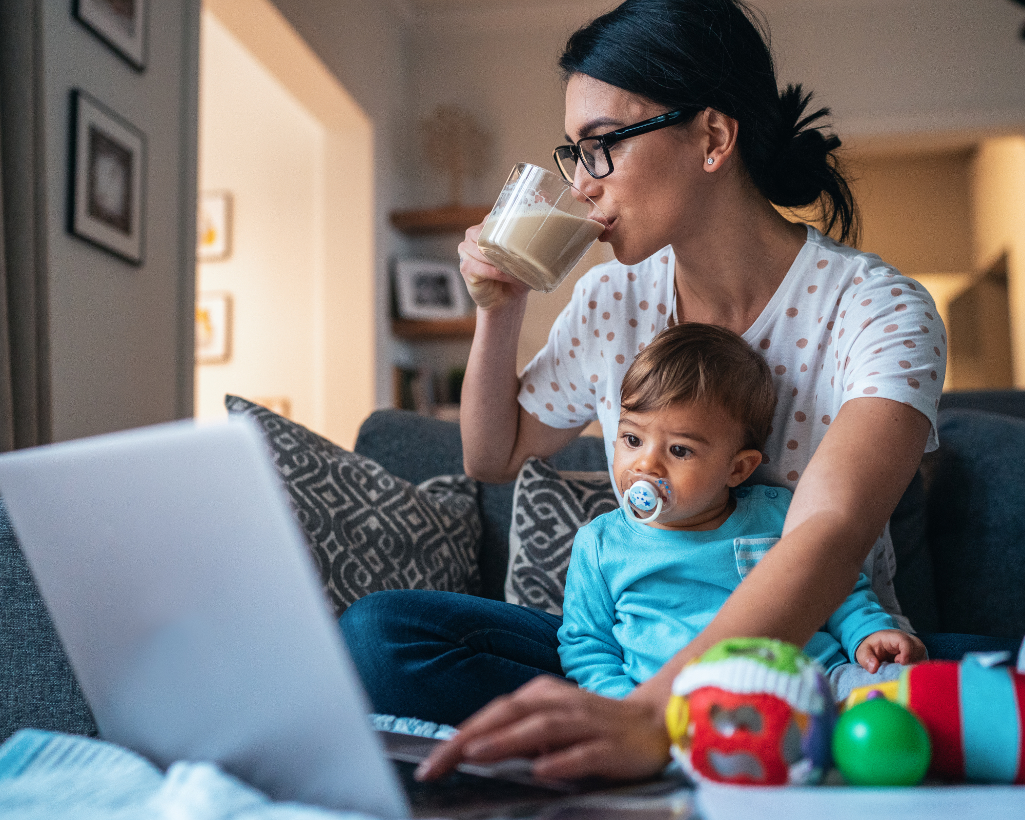 Woman sitting on the floor looking at her laptop and drinking coffee with a child in her lap