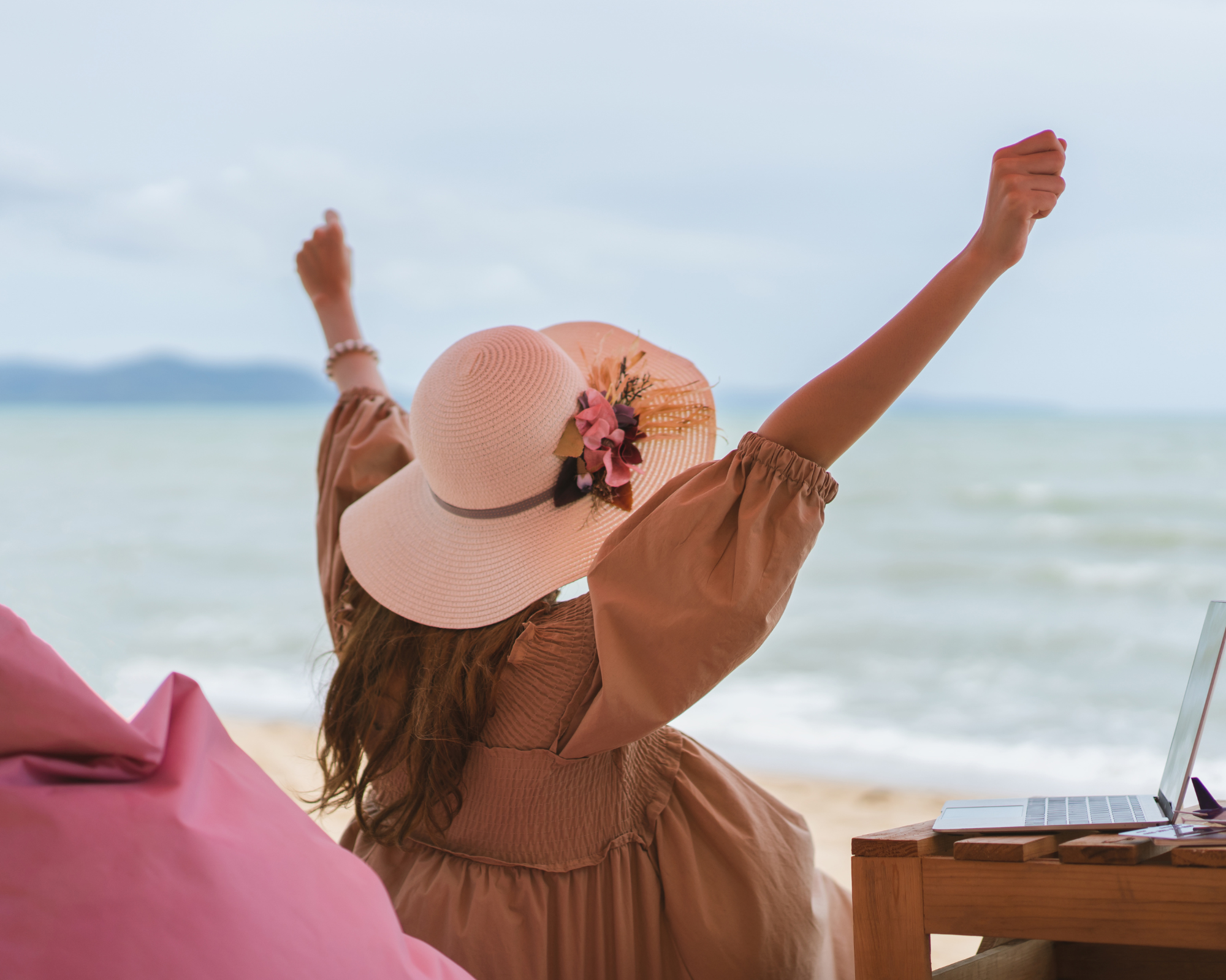 Woman wearing a sun hat sitting on a beach with laptop with arms raised up in praise and the ocean view in the distance- the best remote jobs for stay at home moms