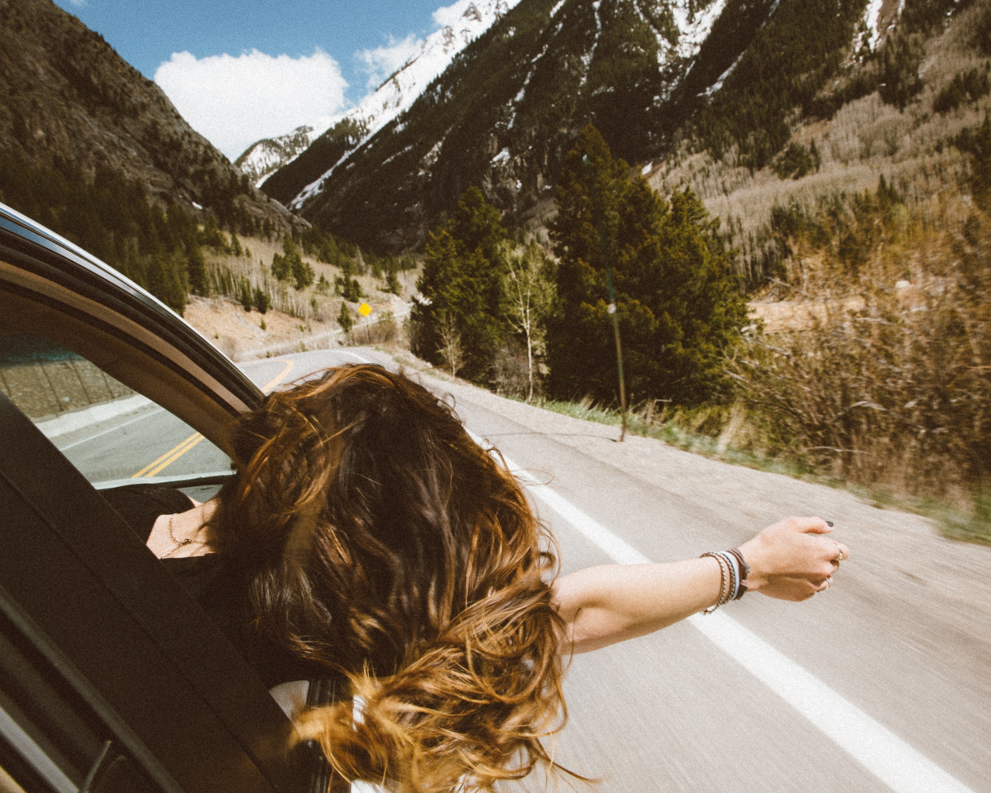 Woman with long hari sticking her head out of a car window during a drive on a road- get paid to travel as a sleep consultant