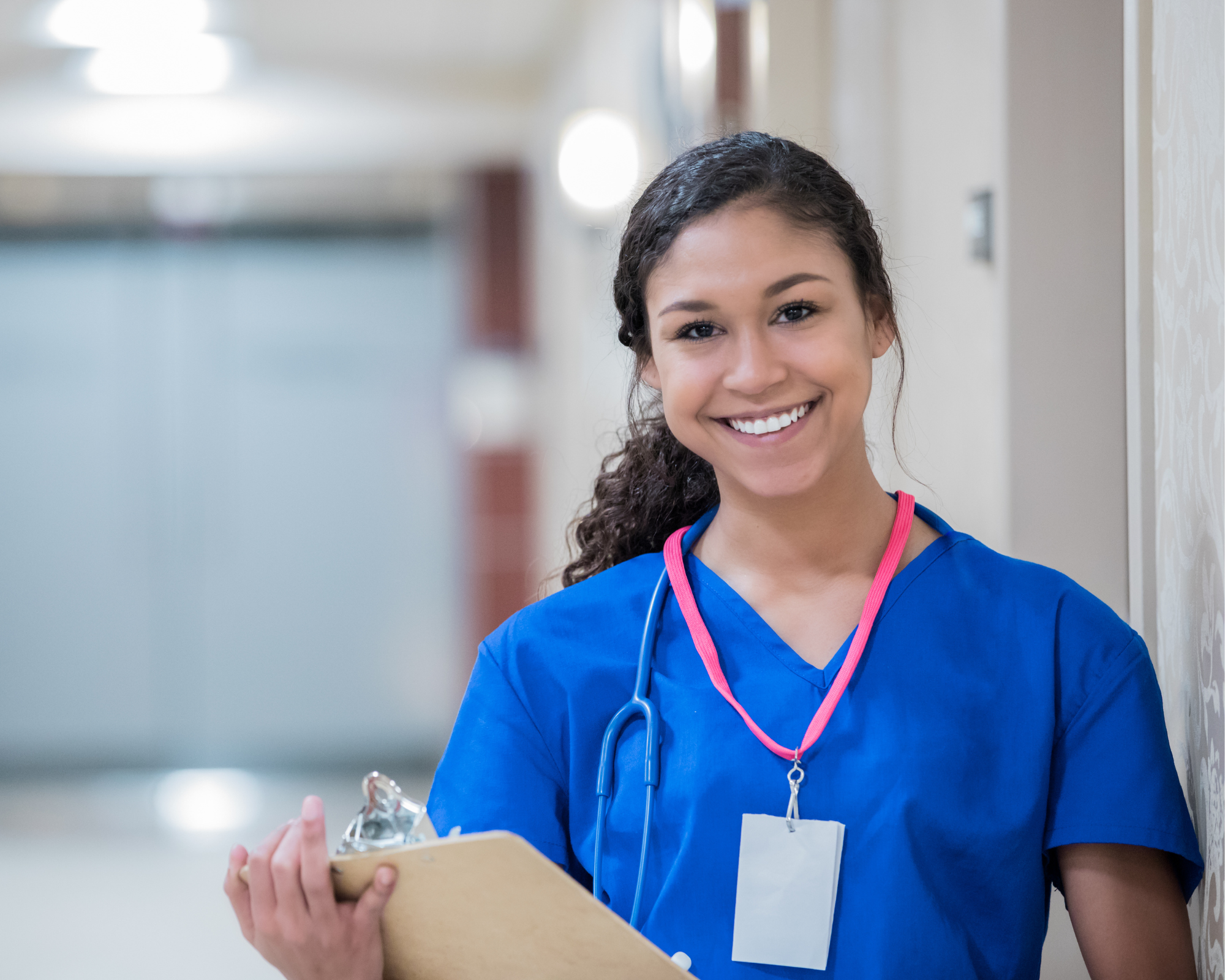 A female nurse wearing blue scrubs and holding a clipboard smiling at the camera.