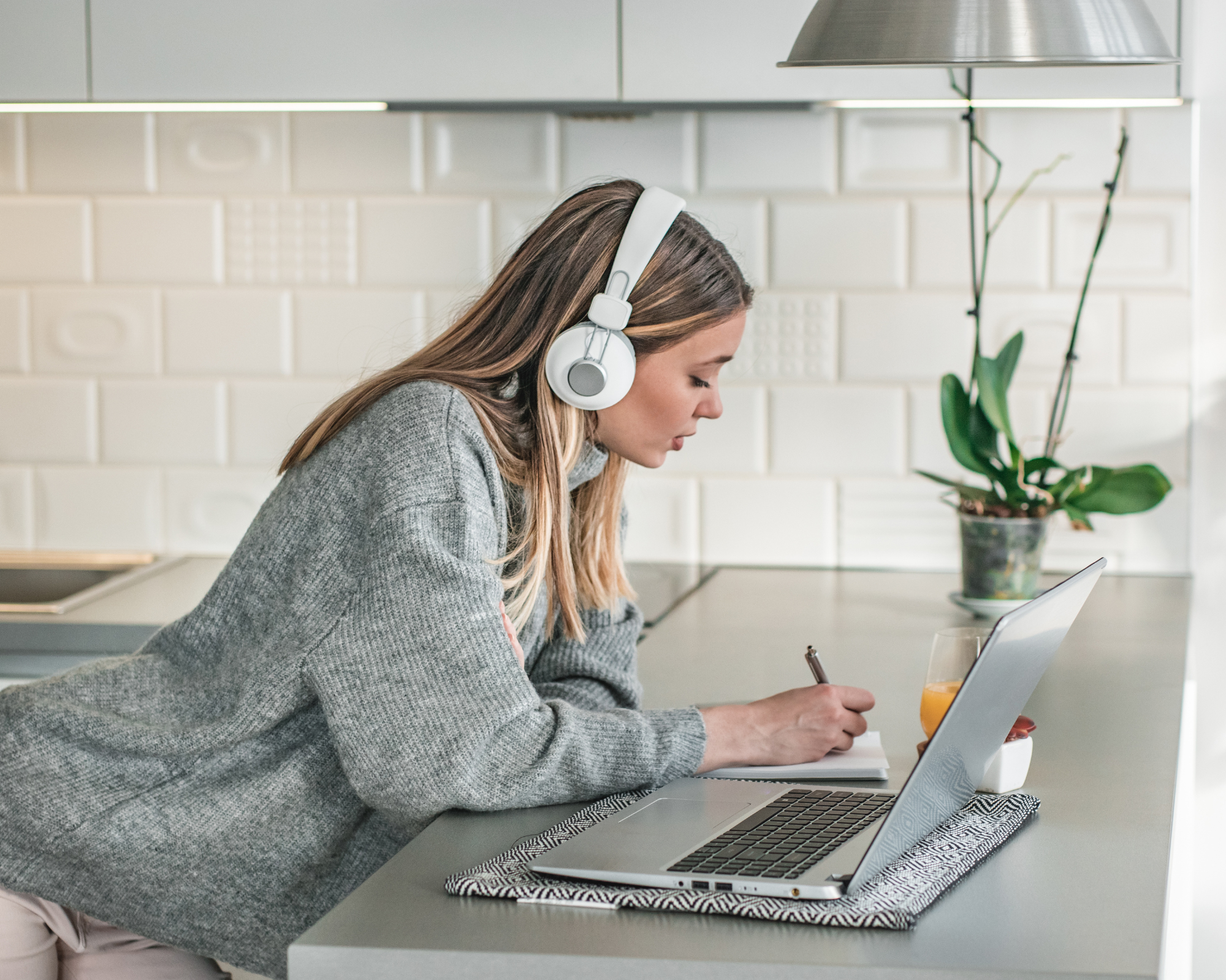 Woman leaning on the counter writing notes with headphones on and an open laptop in front of her. Best online baby sleep training certification